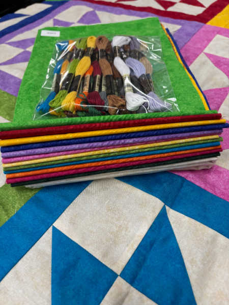 A neatly folded stack of colourful quilting fabrics topped with a clear packet of DMC embroidery threads, part of the Hector’s Year 2026 Block of the Month kit from Patchwork Dreamer.