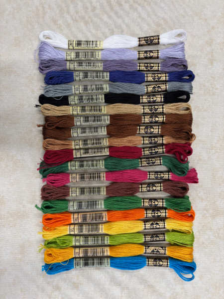 A full selection of DMC embroidery threads in shades of white, purple, blue, brown, green, orange, yellow, and red, neatly arranged on a cream background.