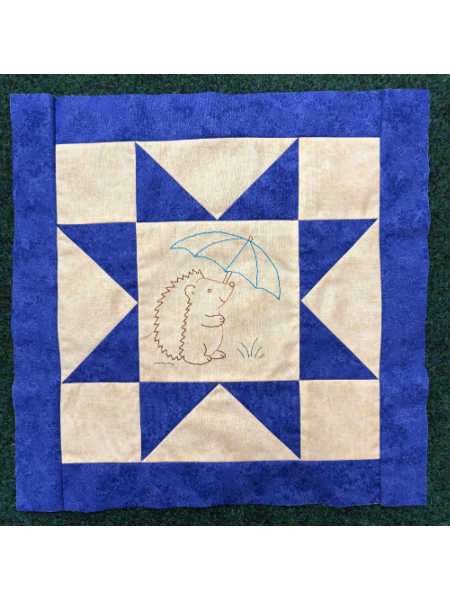 April block from Hector’s Year BOM showing a blue and cream star block with an embroidered hedgehog holding an umbrella.