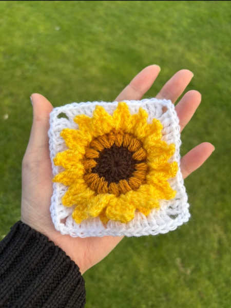 Hand-crocheted sunflower square with raised yellow petals on a white background, held in the palm of a hand outdoors. Crochet class in Bodmin Cornwall UK